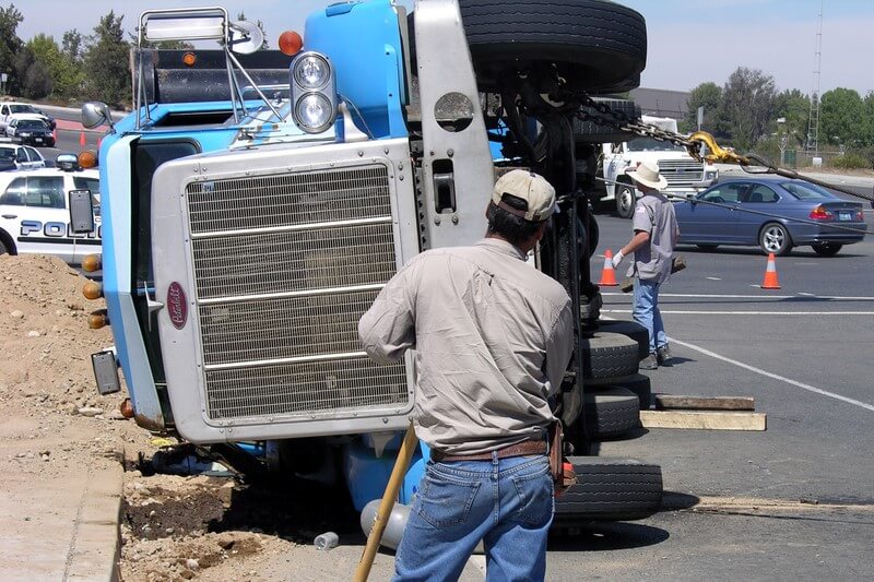 a blue truck has tipped over on the side of the road with a worker in the foreground looking on
